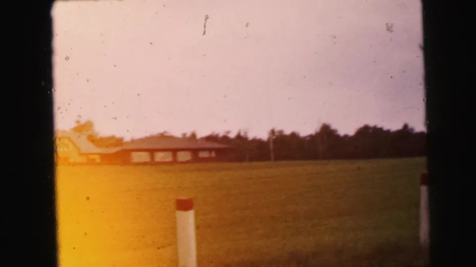 A faded 1964 home-movie frame shows an open field, a low distant building, trees, and white posts near Camden, New Jersey.