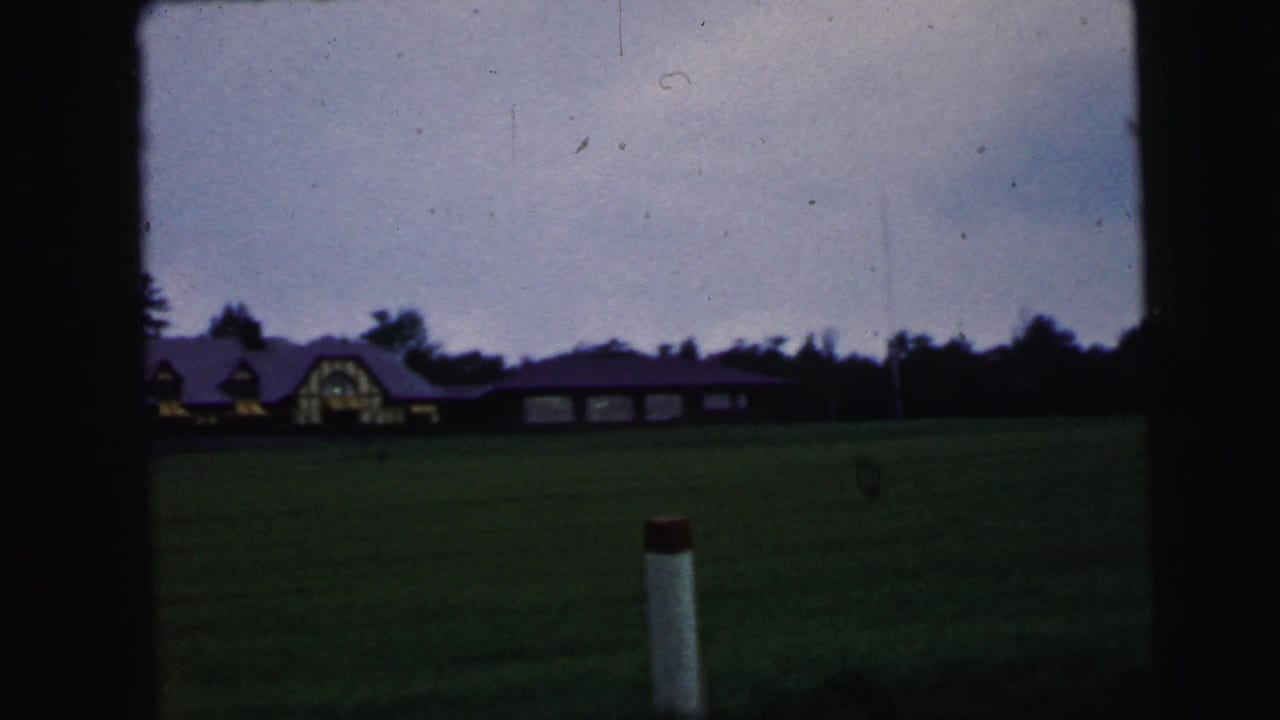 1964 Camden, New Jersey home-movie frame showing a low gabled building across a field beneath a darkening sky.
