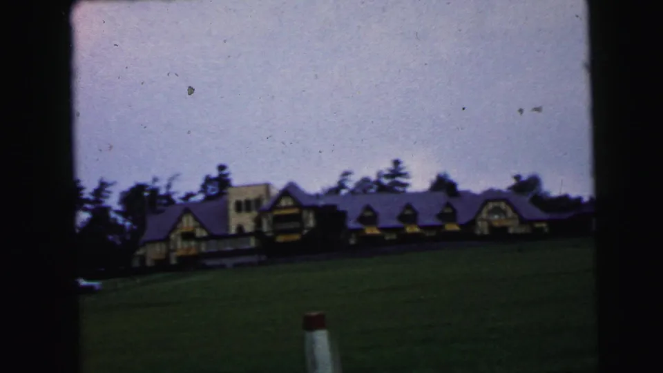 1964 home-movie frame with the building larger in view, trees behind it, and a dark lawn under storm light.