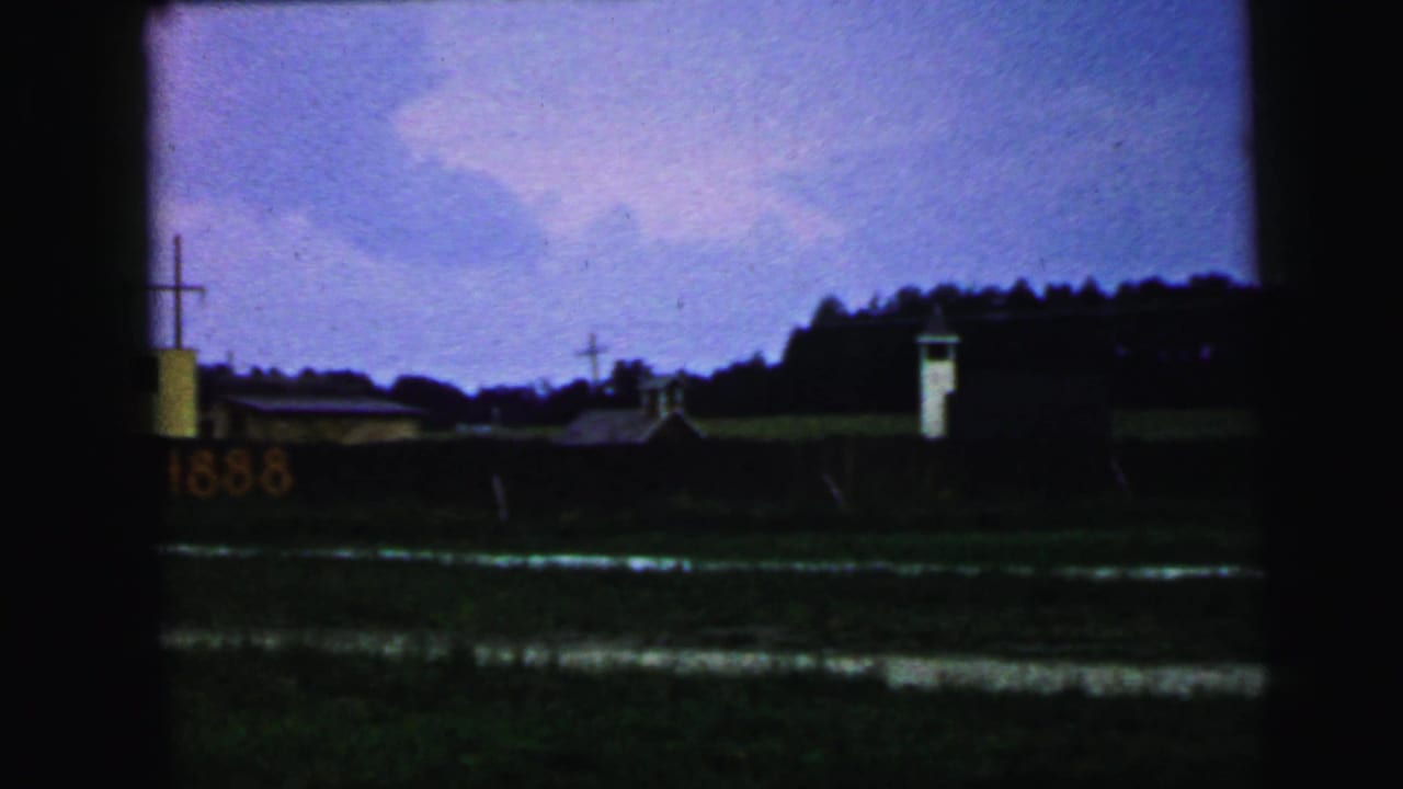 1964 Camden, New Jersey home-movie frame showing a distant yellow building with a cross, a rail car display, a white tower, and dark foreground ground.