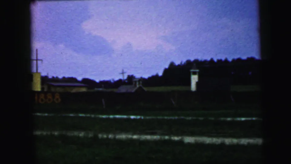 1964 Camden, New Jersey home-movie frame showing a distant yellow building with a cross, a rail car display, a white tower, and dark foreground ground.