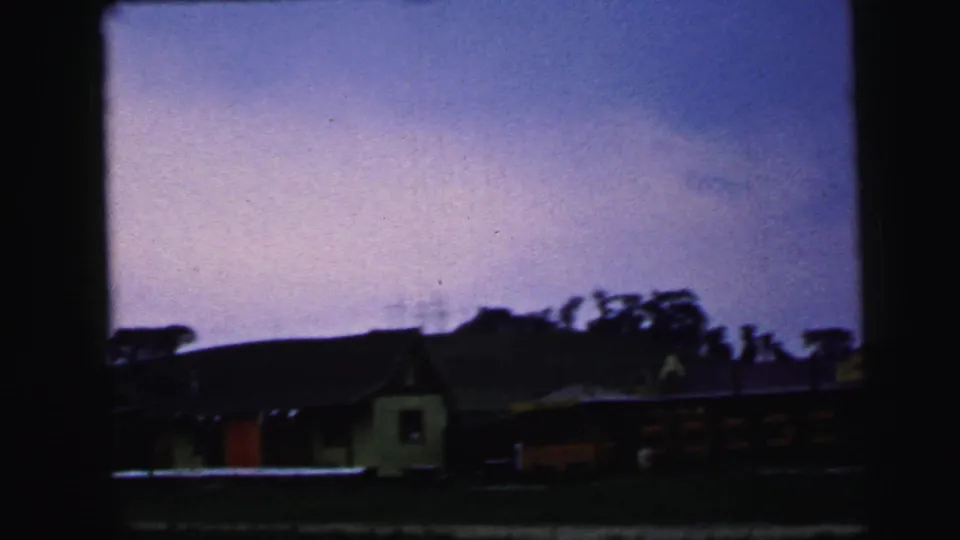 1964 Camden roadside view with a small pale building, red structure, dark roofline, trees, and partial commercial signage passing through frame.