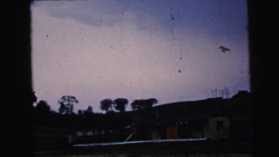 1964 Camden 8mm frame with dark roadside buildings, a small pale building, utility poles, silhouetted trees, and a clouded evening sky.