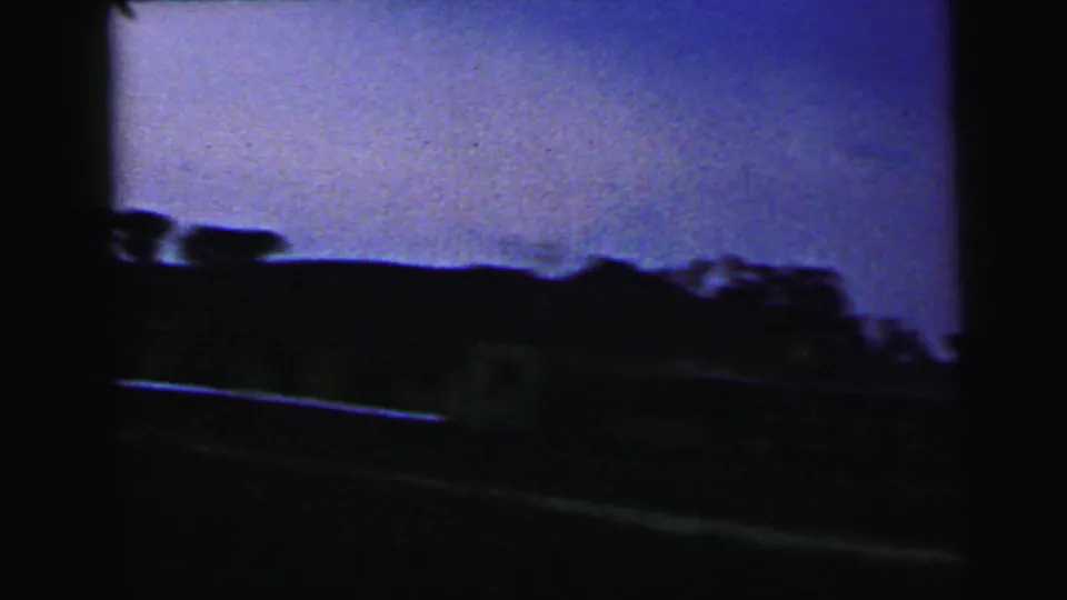 1964 Camden moving-vehicle frame with a blurred dark tree line, low horizon, purple sky, and roadside foreground softened by motion.