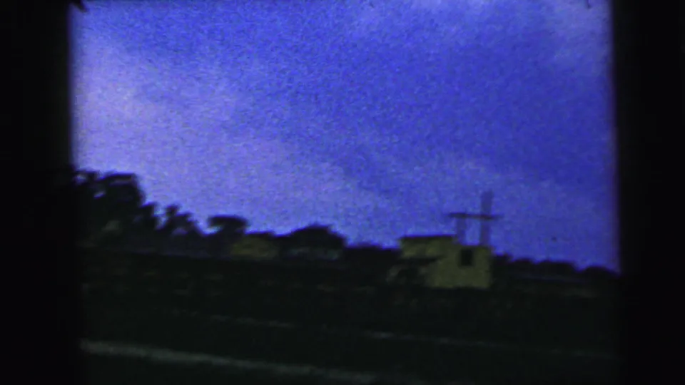 1964 Camden-area 8mm frame showing a dark roadside horizon, trees, a small pale structure, and a utility pole under blue dusk sky.