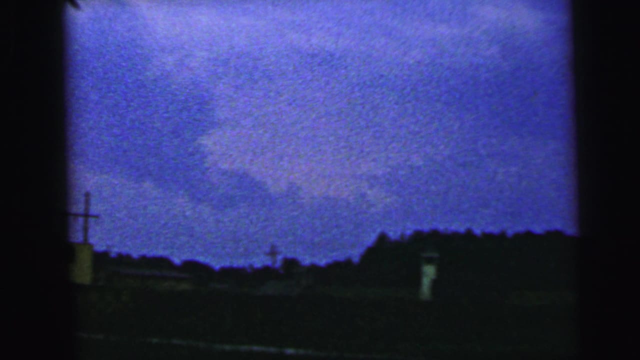 1964 Camden-area home-movie frame with a dark rural horizon, faint low buildings, utility poles, and grainy blue-purple sky.
