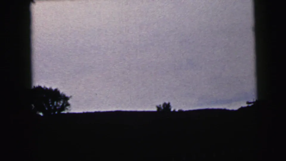 1964 Camden-area 8mm frame showing silhouetted roadside trees and a low dark horizon beneath a pale gray-blue dusk sky.