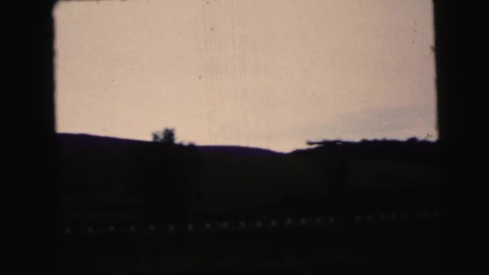 1964 Camden-area 8mm frame with a dark rolling horizon, small tree silhouettes, and warm late-day sky near sunset.