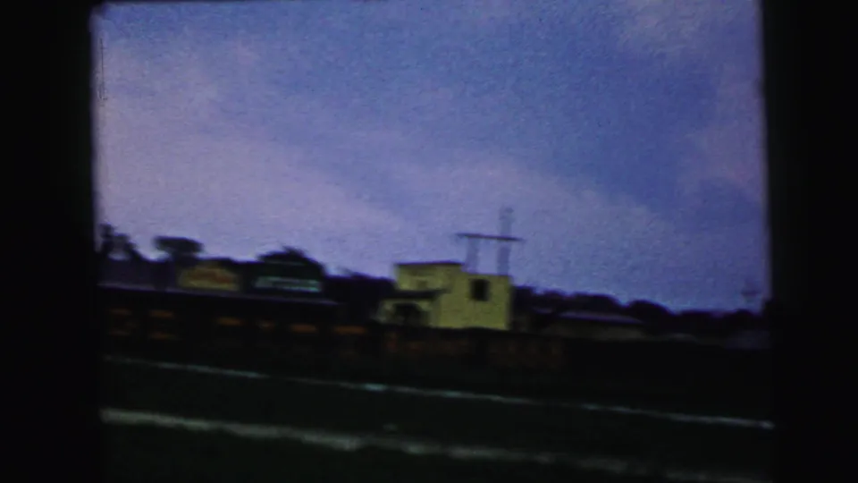In this 1964 8mm Camden-area frame, blurred train cars pass along tracks near low buildings and a distant dark tree line.