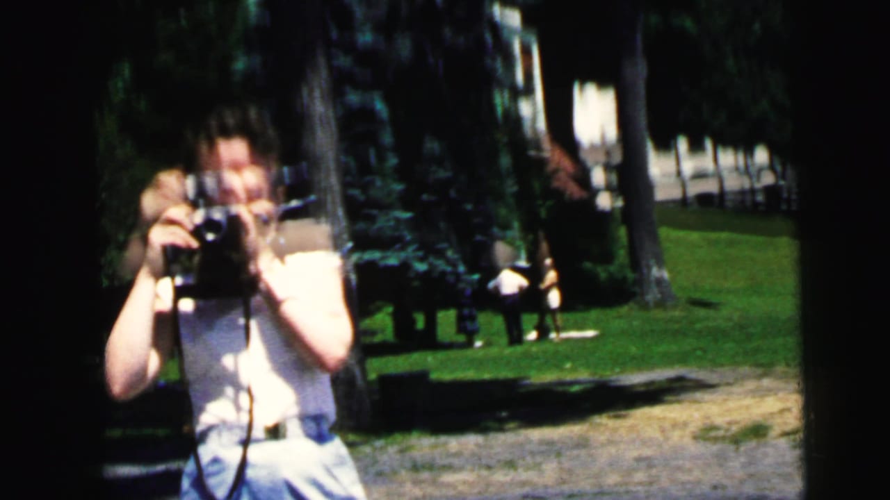 Woman holding an 8mm camera in a sunny park with trees and people in the distance.