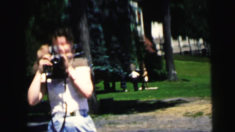A woman in light summer clothing films with an 8mm camera in a Camden, New Jersey park setting, based on supplied 1964 metadata.