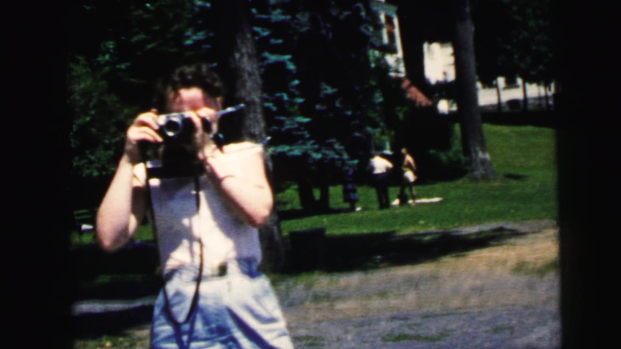 The woman continues filming in a tree-lined park, with distant visitors and grass visible behind her in the 1964 home-movie frame.