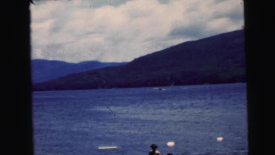 A lake view shows dark mountain slopes, cloudy sky, swimmers, and floating buoys, recorded as part of the supplied 1964 vacation footage.