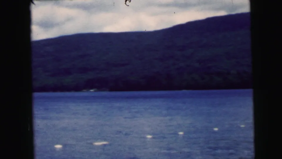 The final frame holds on the lake and mountain backdrop, with small swimmers and buoys visible in the water under a cloudy sky.