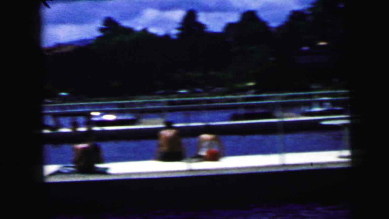 Waterside dock with seated people, water, trees, and a dark foreground edge.