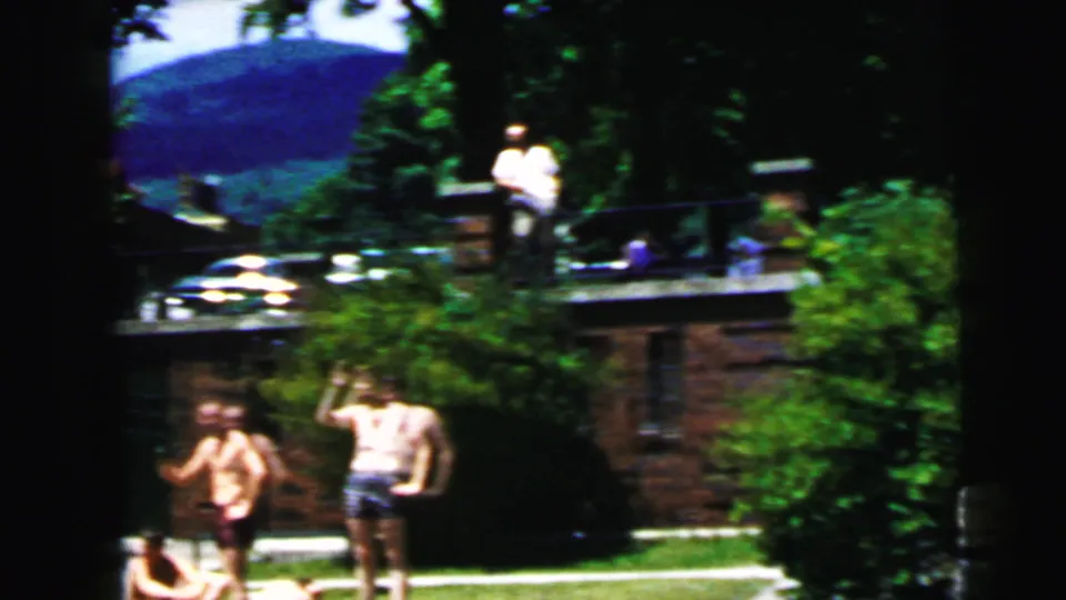 1964 home-movie frame of visitors in the Jolly Roger grounds near a railing, lawn, trees and distant ridge.