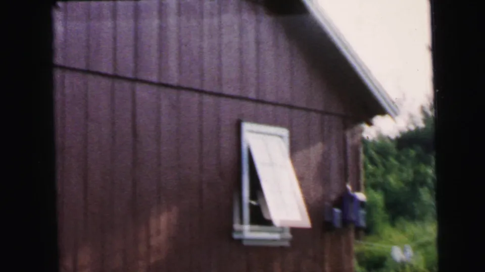 1964 Camden home-movie frame showing a red bungalow side wall, open white window, roof angle, and a small area of greenery.