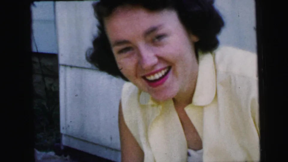 A dark-haired woman in a pale sleeveless dress smiles toward the camera in Camden, New Jersey, on 1964 8mm home-movie film.