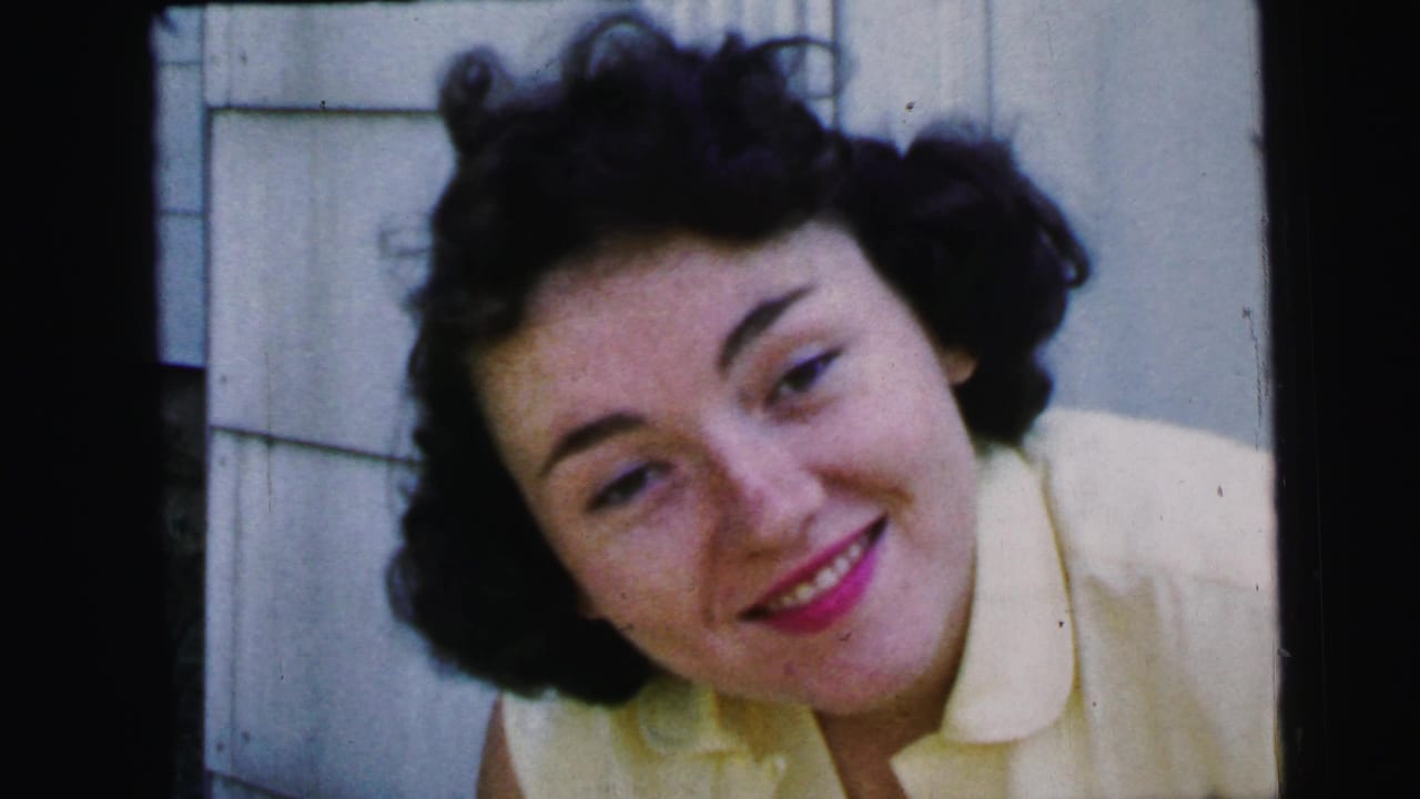 Young woman with dark curled hair smiles beside a white paneled wall.