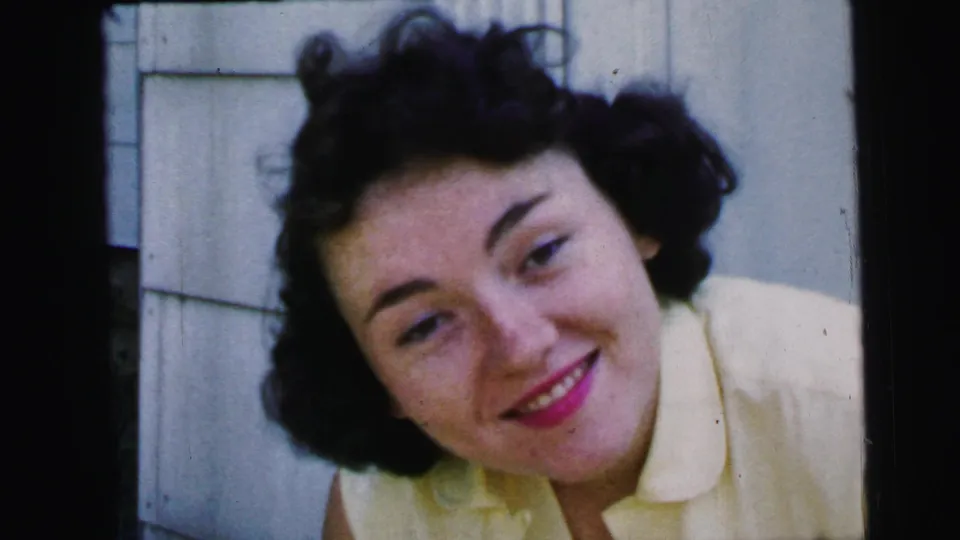 In 1964 Camden home-movie footage, a young woman in a pale yellow collared vest smiles warmly near a white paneled wall.