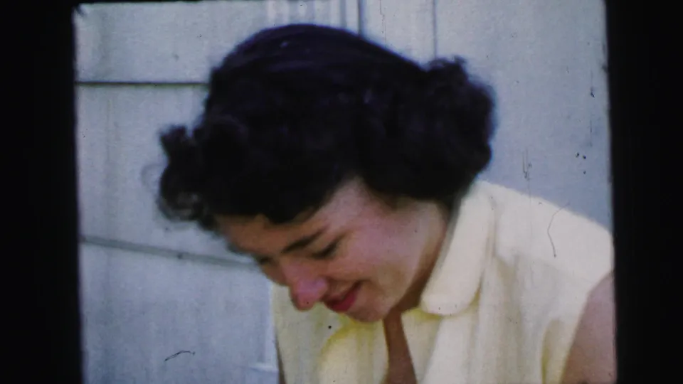 The young woman lowers her gaze with a modest smile, still framed close against the white wall in 1964 Camden footage.