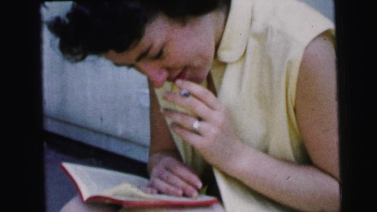 Woman in a yellow sleeveless dress smokes while reading a red-covered book outdoors.