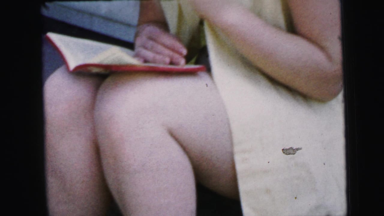 Close 8mm view of the woman's lap, yellow dress, and hands near a red-covered book during the Camden 1964 outdoor scene.