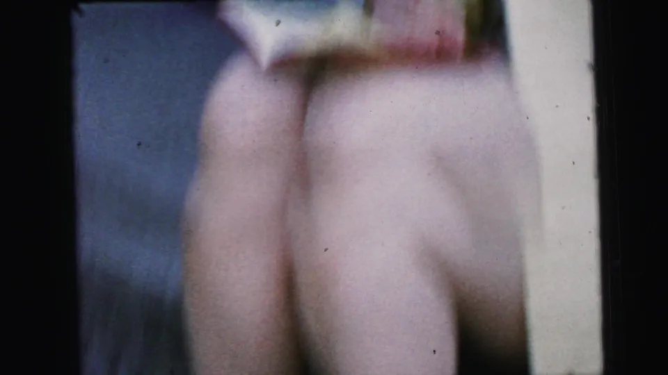 Soft-focus Camden 1964 frame showing the seated woman's legs, flat shoes, yellow dress edge, and blurred red-covered book.