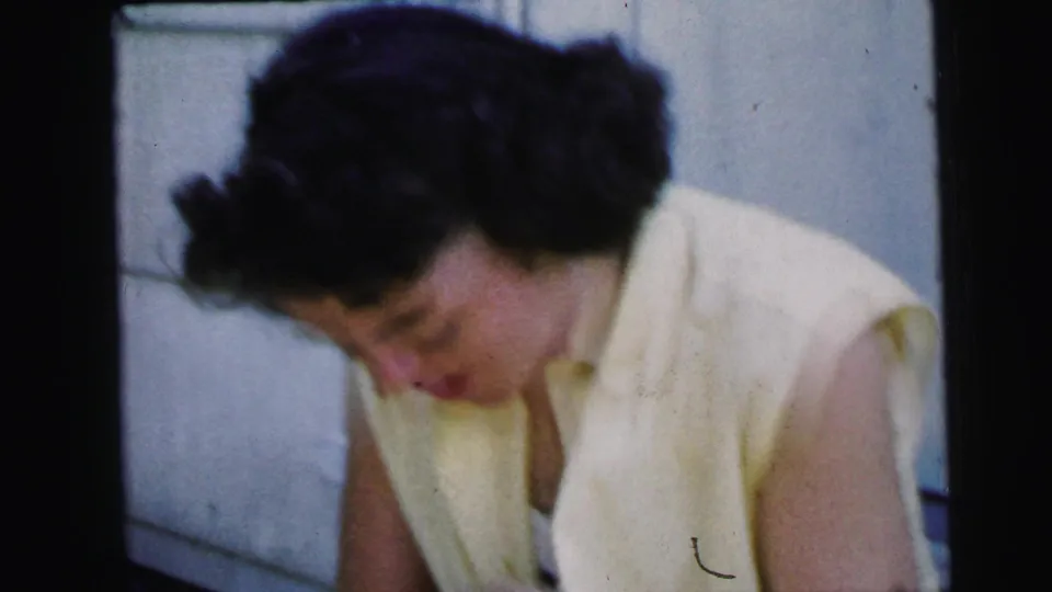 The 1964 Camden home-movie frame shows the woman looking down, her short dark hair and yellow sleeveless dress filling the close composition.
