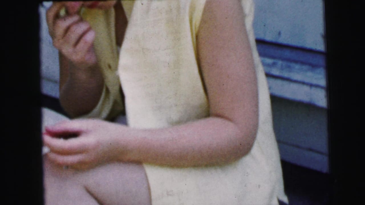 In Camden, New Jersey, 1964 home-movie footage, a seated woman in a pale yellow sleeveless dress holds a cigarette and notepad on a porch step.