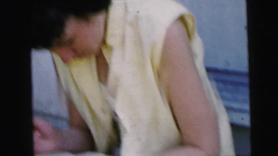 A woman in a pale yellow sleeveless dress leans forward on a Camden porch in 1964, partly blurred by handheld 8mm movement.