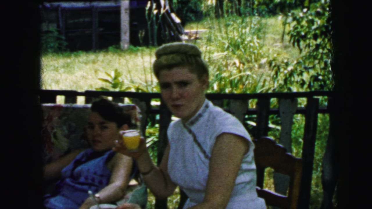 Two women relax on a shaded porch in 1964 Camden footage, with one in a white sleeveless top holding an orange drink.