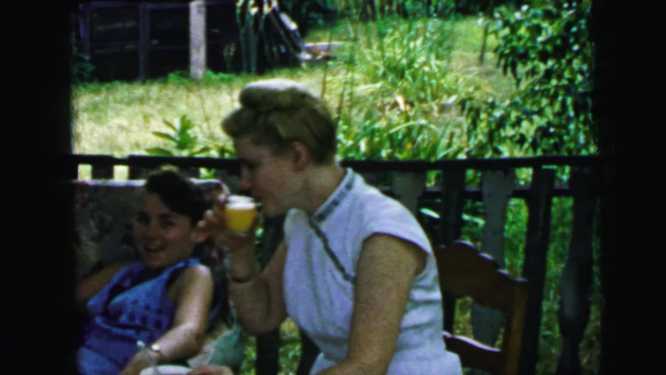 Two women in 1964 Camden home-movie footage sit on a shaded porch, with one holding an orange drink and facing her companion.
