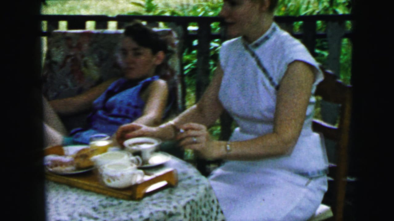 1964 Camden home-movie frame showing women at a small outdoor tea table with cups, saucers, pastries, and greenery behind them.