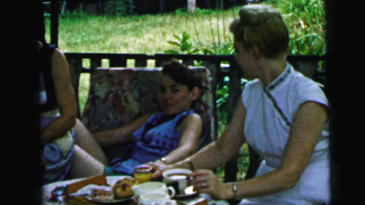1964 Camden garden gathering with women seated near tea cups and pastries, partly blocked by a dark vertical post in the frame.