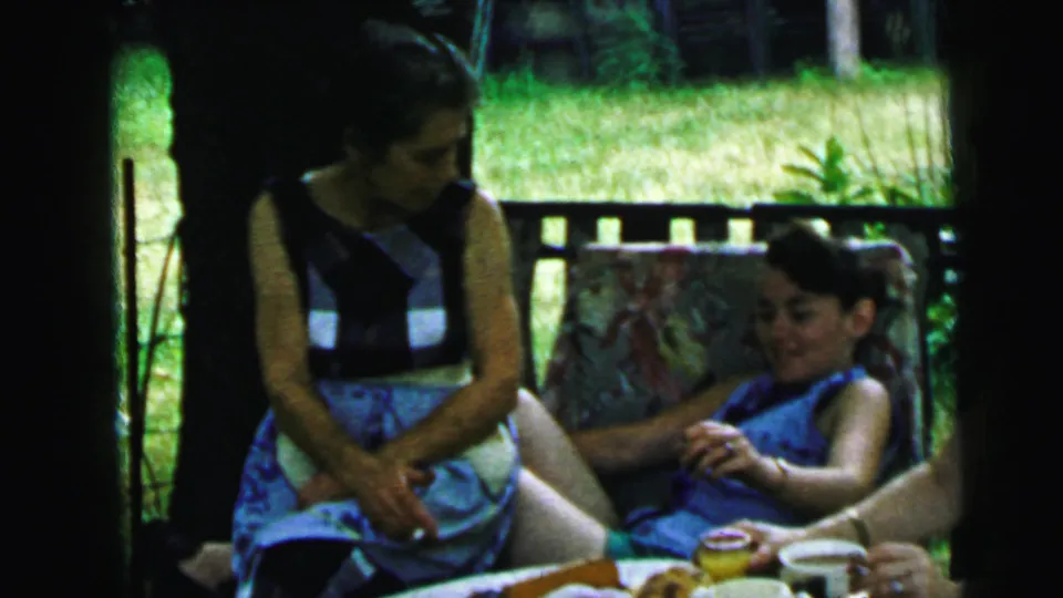1964 Camden home-movie frame of women in blue dresses seated beside an outdoor tea table during a garden gathering.