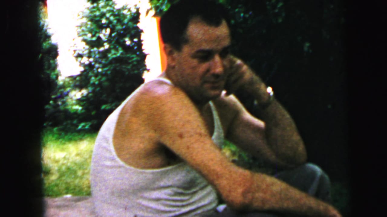 In 1964 Camden home-movie footage, a man in a white tank top sits on stone steps beside grass and shrubs.