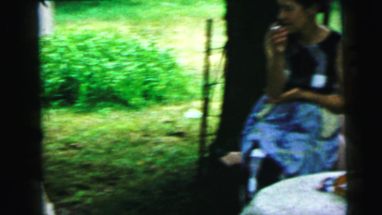 1964 Camden home-movie frame showing adult women seated on a shaded porch, with drinks, summer clothing, and a green yard beyond the railing.