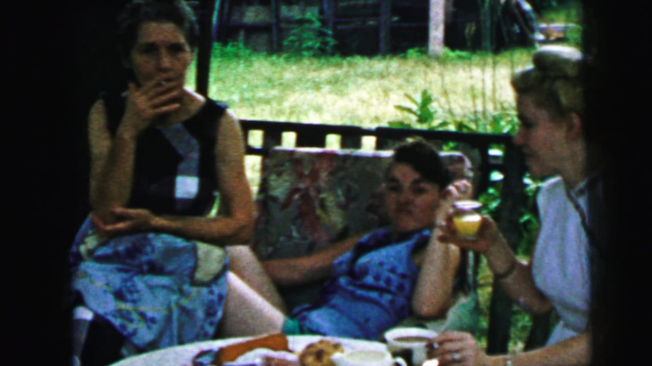 Adult women gather on a porch in Camden, New Jersey, with cups and plates on the table and one woman reclining beside the railing.