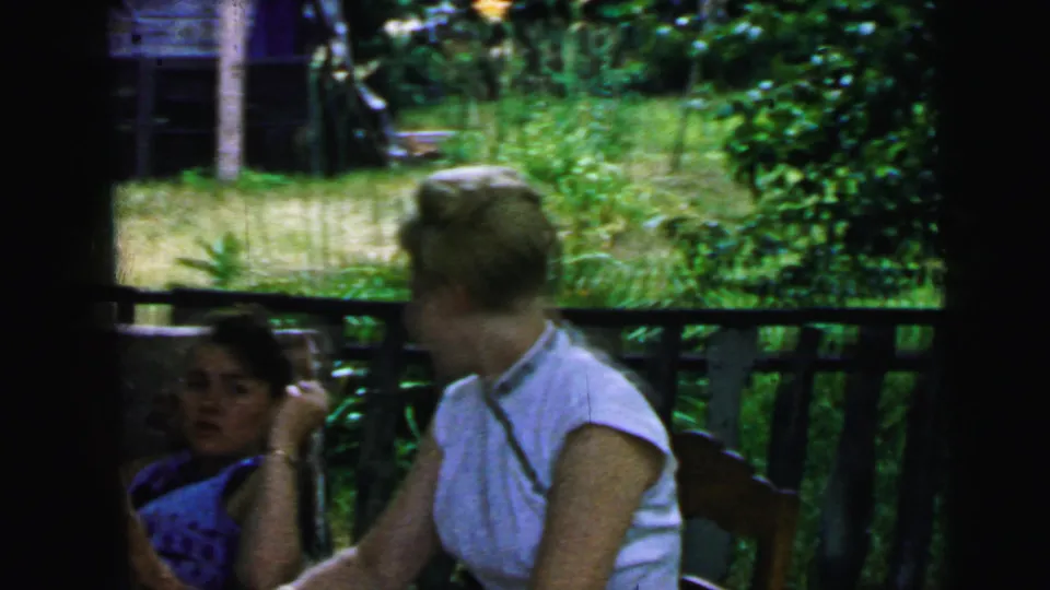 Camden home-movie frame of women on a porch, including a seated woman partly in shadow and another turned toward her near the railing.