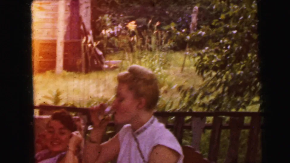 1964 Camden porch scene showing an adult woman near the railing who appears to smoke, with warm light leak and foliage in the yard behind her.