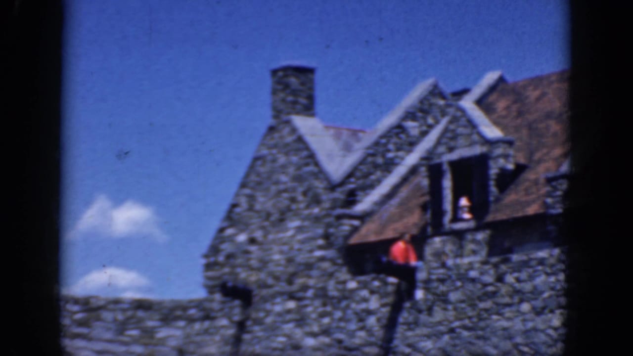 Stone castle-like building with dormer windows, chimney, and visitors near a low wall.