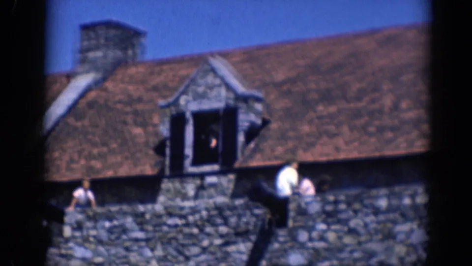 1964 8mm frame with visitors beside a stone wall below a dormer window on a castle-like building in Charlestown, North Carolina.