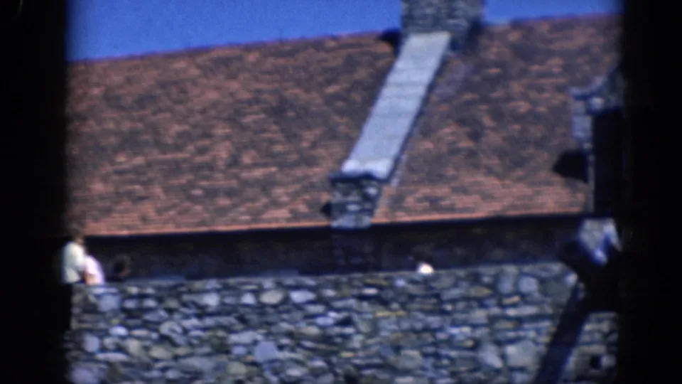 1964 home-movie frame focusing on the roof and stone wall of a castle-like structure, with visitors partly visible at the edge.