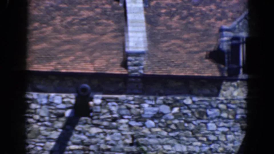 1964 8mm frame of a stone landmark roof with chimney, dormer, cannon, and battlement wall in bright sunlight.