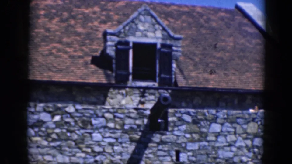 Final 1964 Charlestown frame showing the stone building roof, open dormer, cannon on the wall, and a distant visitor.