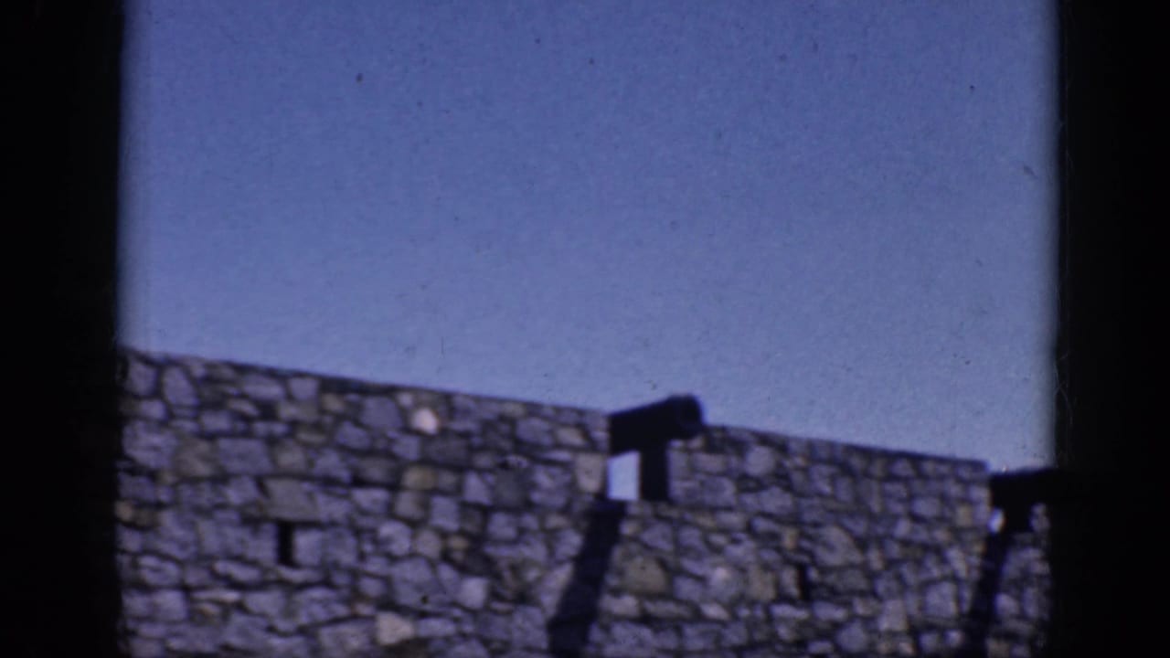 1964 8mm frame from Charlestown, North Carolina, showing a rough stone wall, blue sky, and distant people along the upper edge.