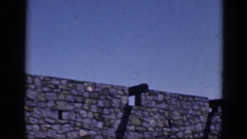 1964 8mm frame from Charlestown, North Carolina, showing a rough stone wall, blue sky, and distant people along the upper edge.