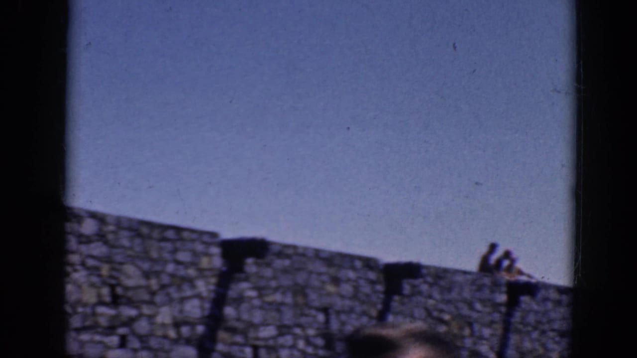 1964 8mm frame showing the stone wall and sky, with a partial person visible at the lower right edge.
