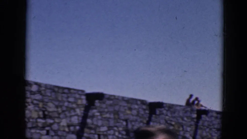 1964 8mm frame showing the stone wall and sky, with a partial person visible at the lower right edge.